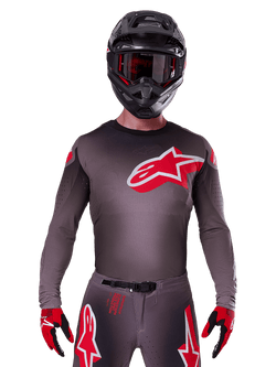 Alpinestars 2025 Supertech Lipan Jersey, motocrosströja, smoke och röd, med en stor röd och vit logotyp på bröstet, bärs med en svart hjälm och röda och svarta handskar, atletisk racingpassform med laserskurna perforeringar