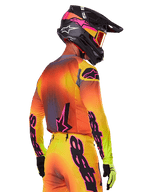 Motocrossförare sedd snett bakifrån iklädd Alpinestars 2025 Supertech Lipan Gear Set, med en färgtoning i gult, orange och rött med svarta och rosa logotyper, svart hjälm med rosa detaljer och orangea goggles, professionell utrustning för terrängkörning.