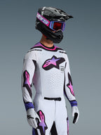 Motocrossförare i Alpinestars 2026 Supertech AFD Vista-utrustningsset, vit tröja och byxor med stjärnlogotyper i rosa och lila gradient, svart hjälm med lila detaljer, lila goggles med rödtonade linser och matchande vita handskar, professionell outfit för terrängkörning
