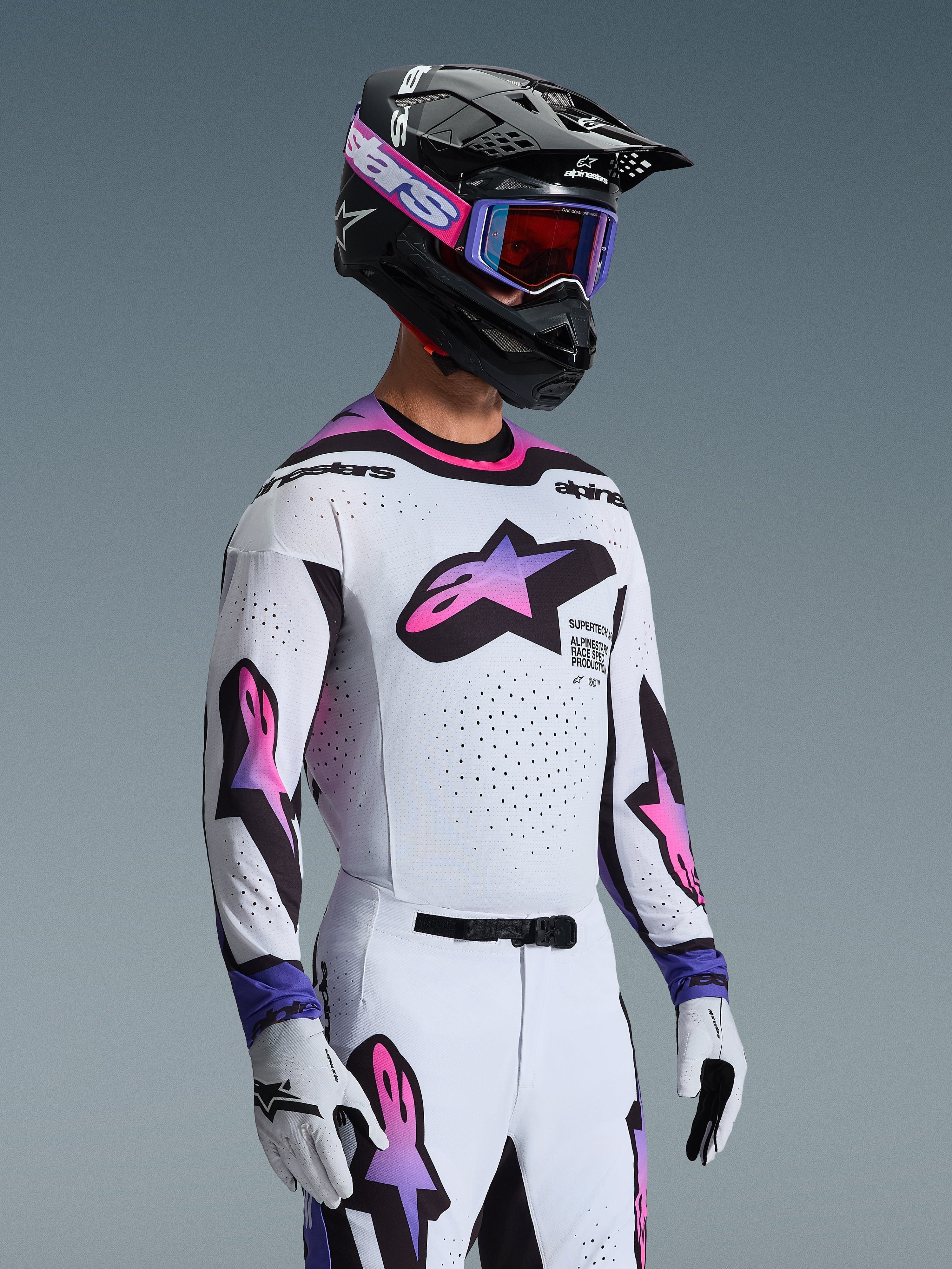 Motocrossförare i Alpinestars 2026 Supertech AFD Vista-utrustningsset, vit tröja och byxor med stjärnlogotyper i rosa och lila gradient, svart hjälm med lila detaljer, lila goggles med rödtonade linser och matchande vita handskar, professionell outfit för terrängkörning