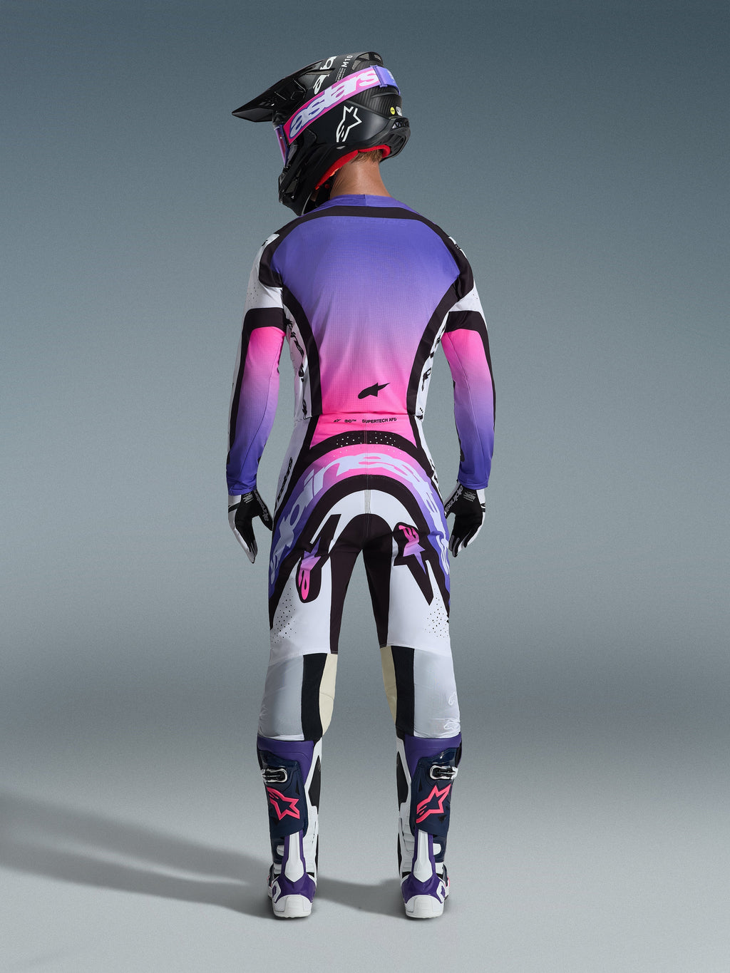 Motocrossförare sedd bakifrån iklädd Alpinestars 2026 Supertech AFD Vista utrustningsset, tröja och byxor med en livfull färggradient i vitt, lila och rosa med svarta detaljer, svart hjälm med lila goggles, matchande lila och vita stövlar, koordinerad professionell racingoutfit för terrängprestanda