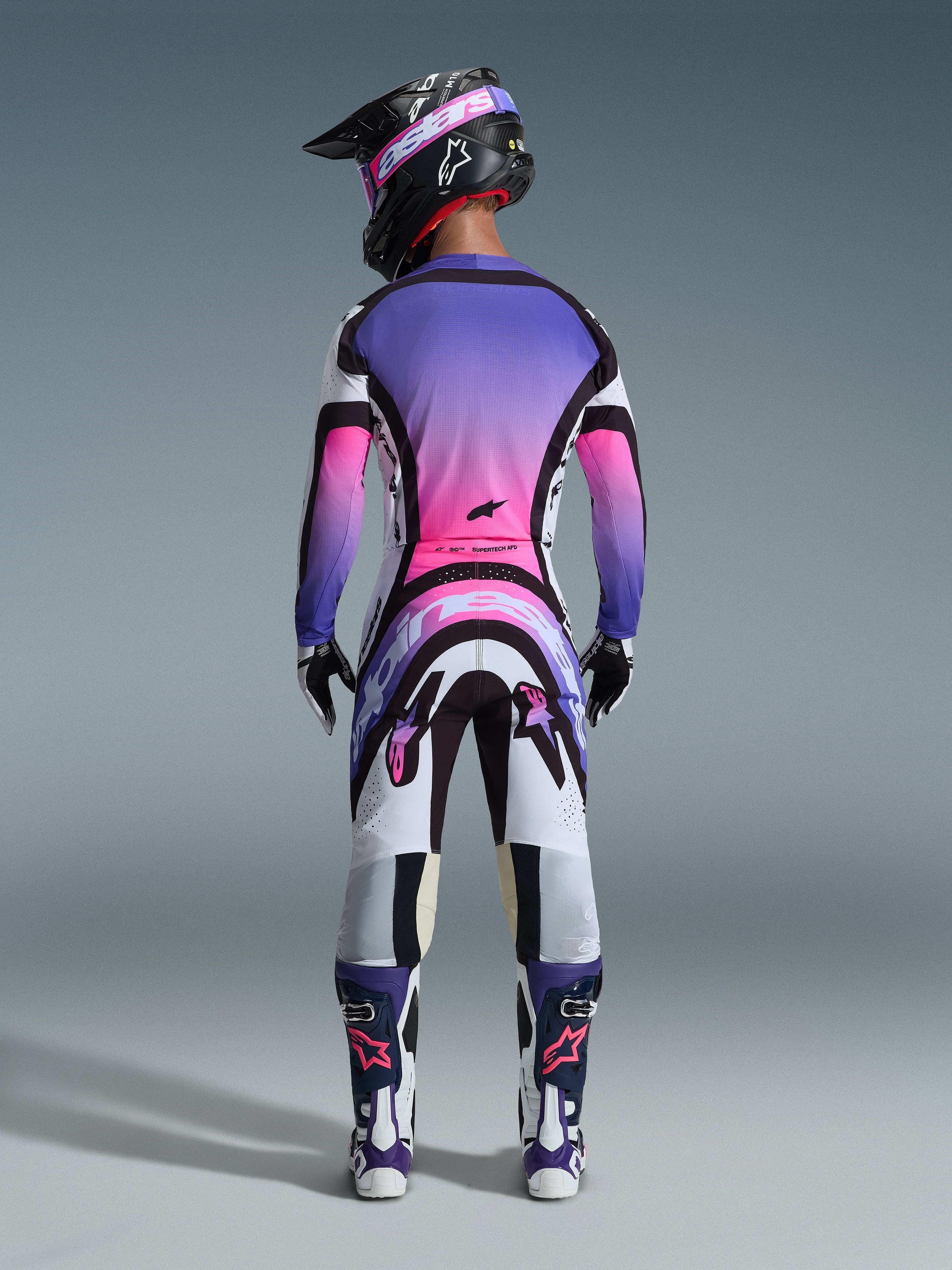 Motocrossförare sedd bakifrån iklädd Alpinestars 2026 Supertech AFD Vista utrustningsset, tröja och byxor med en livfull färggradient i vitt, lila och rosa med svarta detaljer, svart hjälm med lila goggles, matchande lila och vita stövlar, koordinerad professionell racingoutfit för terrängprestanda