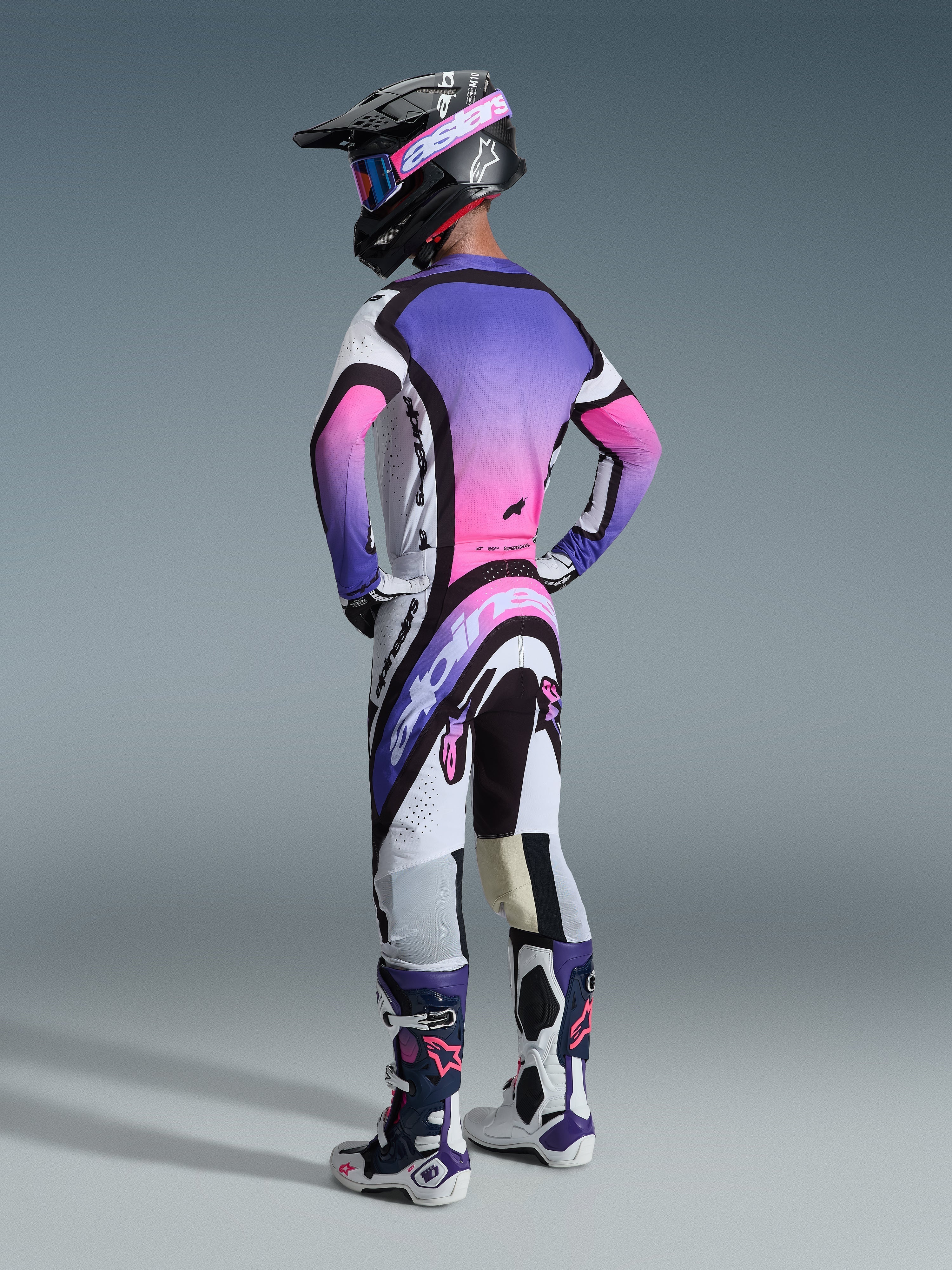 Motocrossförare sedd bakifrån klädd i Alpinestars 2026 Supertech AFD Vista utrustningsset i White Purple Black, med en tröja som har en livfull lila och rosa gradient på ryggpanelen, matchande byxor med tydlig Alpinestars-logotyp, svart hjälm med rosa goggles och matchande vita och lila racingstövlar.