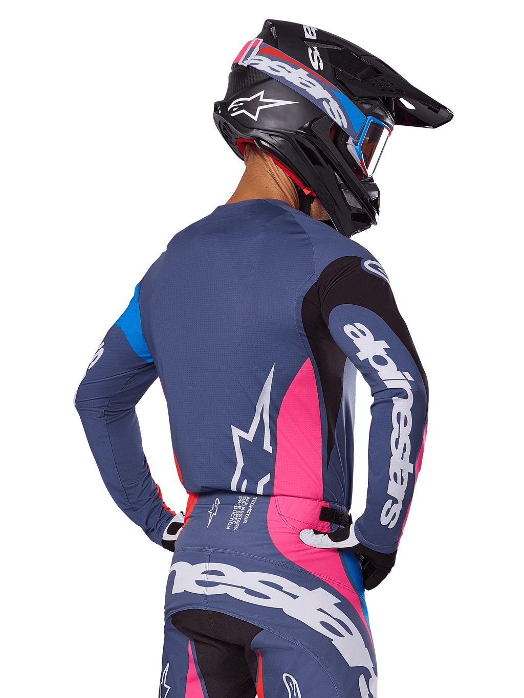 Motocrossförare sedd bakifrån iklädd Alpinestars 2025 Techstar Dreem-utrustningsset i grått, bestående av en blågrå tröja och byxor med rosa och svarta paneler, vita Alpinestars-logotyper på ärmarna och ryggen samt en matchande svart hjälm med blå och rosa grafik.
