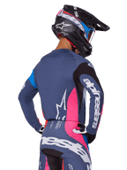 Motocrossförare sedd bakifrån iklädd Alpinestars 2025 Techstar Dreem-utrustningsset i grått, bestående av en blågrå tröja och byxor med rosa och svarta paneler, vita Alpinestars-logotyper på ärmarna och ryggen samt en matchande svart hjälm med blå och rosa grafik.