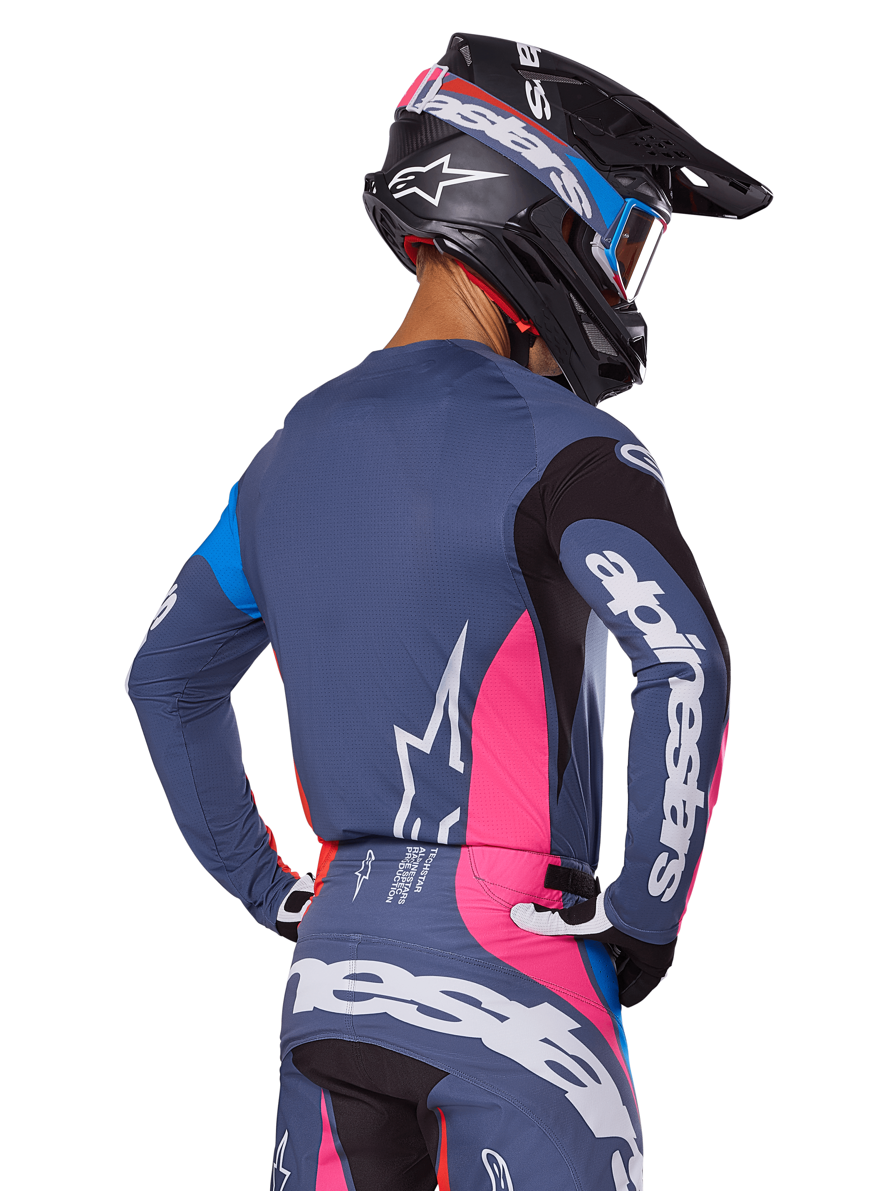 Motocrossförare sedd bakifrån iklädd Alpinestars 2025 Techstar Dreem-utrustningsset i grått, bestående av en blågrå tröja och byxor med rosa och svarta paneler, vita Alpinestars-logotyper på ärmarna och ryggen samt en matchande svart hjälm med blå och rosa grafik.