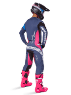 Motocrossförare sedd bakifrån, bär Alpinestars 2025 Techstar Dreem-utrustningsset, grått med livfulla rosa och vita detaljer, svart och vit hjälm och klarrosa stövlar, komplett tävlingsoutfit för motocrossprestanda.