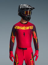 Alpinestars 2026 Supertech Scenz Jersey, motocrosströja, röd, svart och gul, bärs av en förare med matchande svart hjälm och röda handskar, med en tydlig gul 'astars'-logotyp på bröstet och ett svart gradient-spräckligt mönster över axlarna.