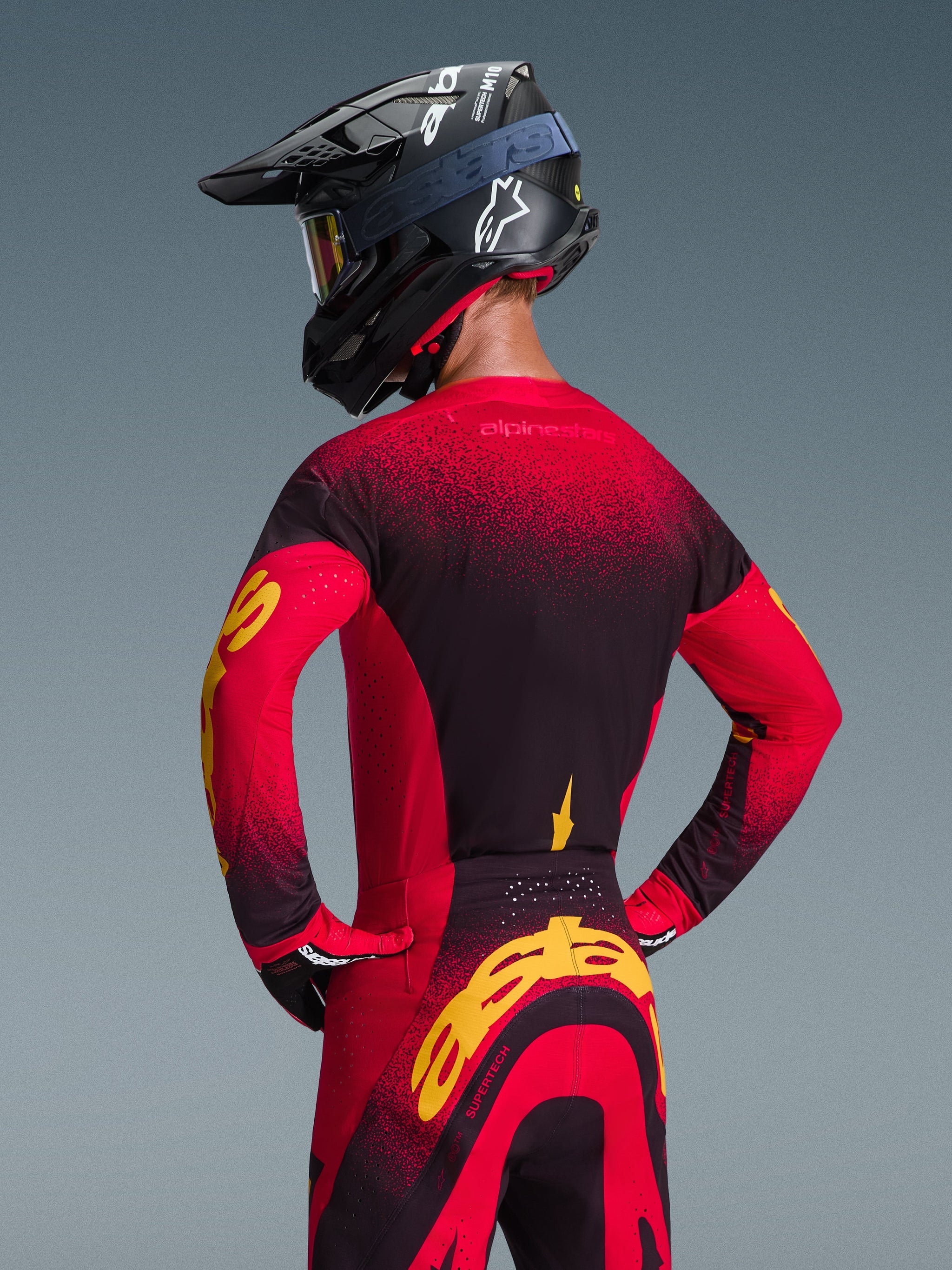 Alpinestars 2026 Supertech Scenz-tröja, röd, svart och gul, ryggvy av en motocrossförare i matchande racingutrustning inklusive terrängbyxor och en svart hjälm, med en röd och svart gradientdesign och framträdande gul Alpinestars-logotyp