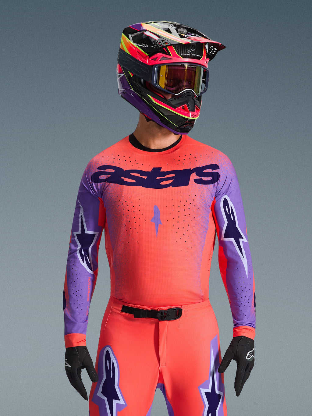 Motocrossförare som bär Alpinestars 2026 Supertech Scenz-utrustningsset, livfull rosa-orange tröja med lila ärmar och 'astars'-branding, matchande byxor med lila stjärndetaljer, flerfärgad racinghjälm med goggles, svarta handskar, professionell off-road racingutrustning