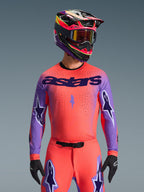 Motocrossförare som bär Alpinestars 2026 Supertech Scenz-utrustningsset, livfull rosa-orange tröja med lila ärmar och 'astars'-branding, matchande byxor med lila stjärndetaljer, flerfärgad racinghjälm med goggles, svarta handskar, professionell off-road racingutrustning