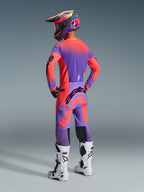 Motocrossförare sedd bakifrån i Alpinestars 2026 Supertech Scenz-utrustningsset, lila och korallrosa tröja och byxor, stor Alpinestars-logotyp på sätet och ärmarna, matchande lila och korallfärgad hjälm, vita och svarta stövlar, koordinerad professionell racingoutfit