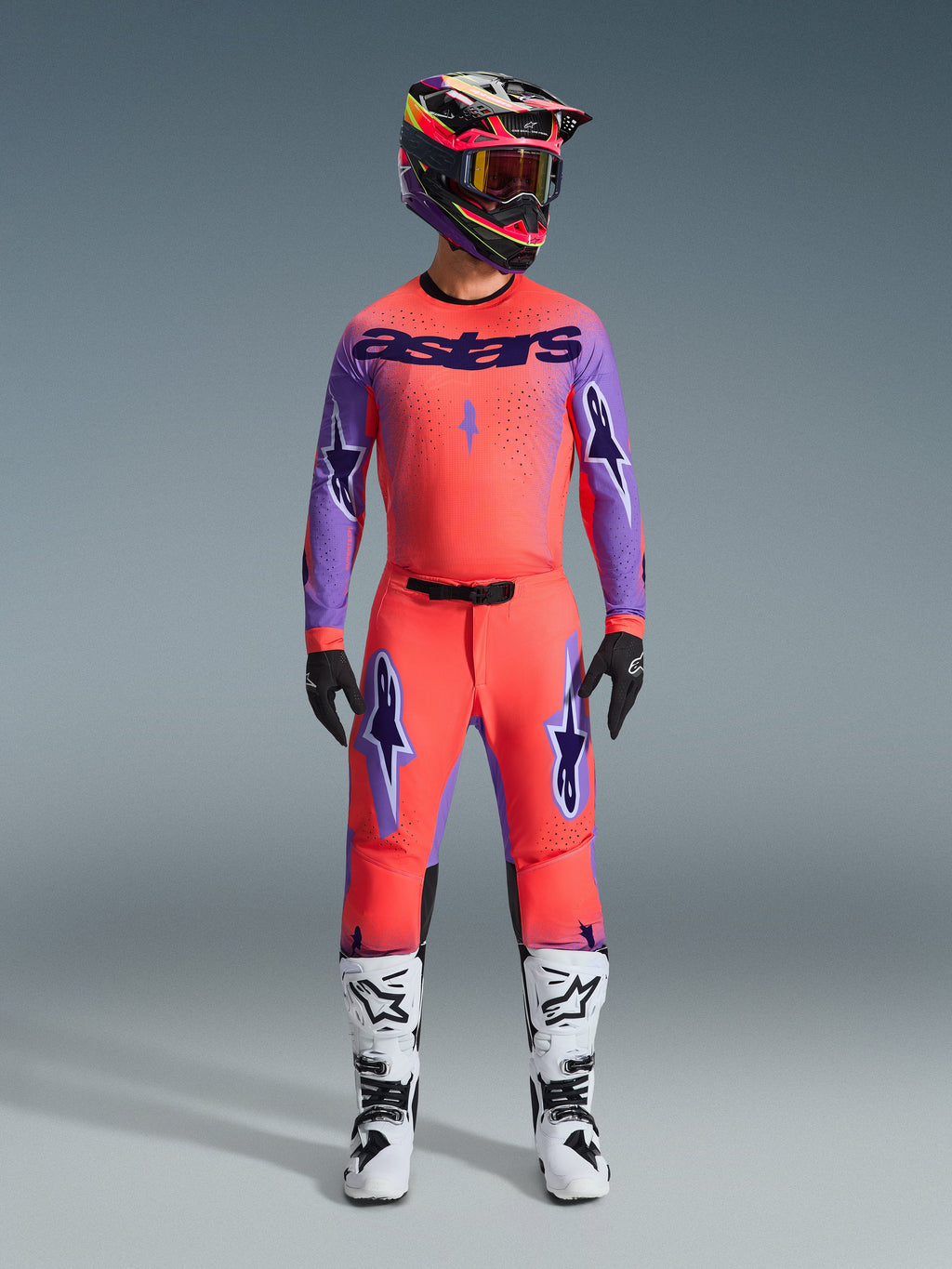 2026 Alpinestars Supertech Scenz utrustningsset, motocrossoutfit i livfull röd-orange och lila, tröja med svart 'astars'-logotyp, matchande byxor med lila stjärnlogotyper, färgstark racinghjälm och vita stövlar, professionell terrängutrustning