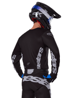 Motocrossförare i Alpinestars 2025 Racer Graphite-utrustningsset, sedd bakifrån, svart tröja och byxor med grå vertikala Alpinestars-logotyper, svart hjälm med blå och vita detaljer samt svarta och blå handskar, koordinerad motocross-racingklädsel