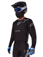Motocrossförare i Alpinestars 2025 Racer Graphite-utrustningsset, svart tröja med grå Alpinestars-logotyper, matchande svarta byxor med grå detaljer, svart hjälm med blå och vita goggles, blå och grå handskar, teknisk outfit för terrängkörning