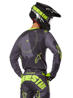 Motocrossförare sedd bakifrån i 2025 Racer Hollow Gear Set, grå tröja med fluorescerande gul cirkulär text och stjärnlogotyper, matchande grå och gula byxor med tydlig logotyp över sätet, svart och limegrön hjälm, koordinerad outfit för terrängkörning