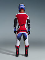 Motocrossförare i Alpinestars 2026 Techstar Knif-utrustningsset, sedd bakifrån, röd, vit och svart tröja med blåtonade detaljer på ärmarna, matchande vita och svarta byxor med framträdande röd branding, blå hjälm och svarta och röda stövlar, professionell racingoutfit för terrängkörning.