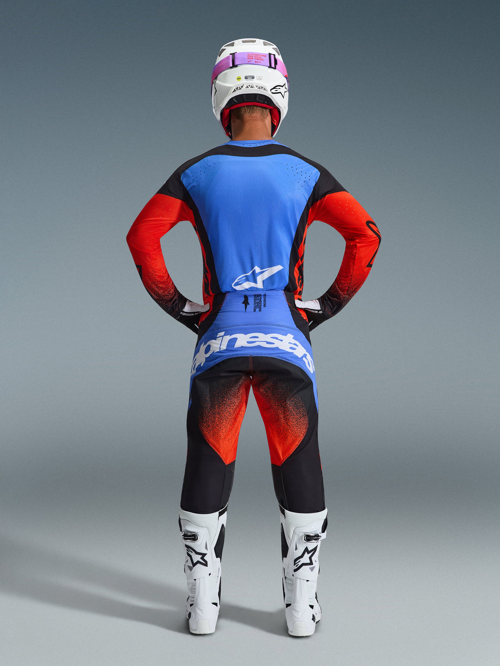 Rear view of a motocross rider wearing the Alpinestars 2026 Techstar Knif Gear Set, blue and red jersey with black accents, black pants with red gradient on the thighs and blue branding, paired with white racing boots and a white helmet with pink goggles.