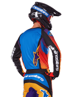 Motocrossförare sedd bakifrån som bär Alpinestars 2025 Fluid Wurx-utrustningsset, flerfärgad tröja med blå rygg och svarta ärmar med vita logotyper, matchande orangea och blå byxor, svart och blå hjälm med orangea goggles, koordinerad racingoutfit för terrängkörning.