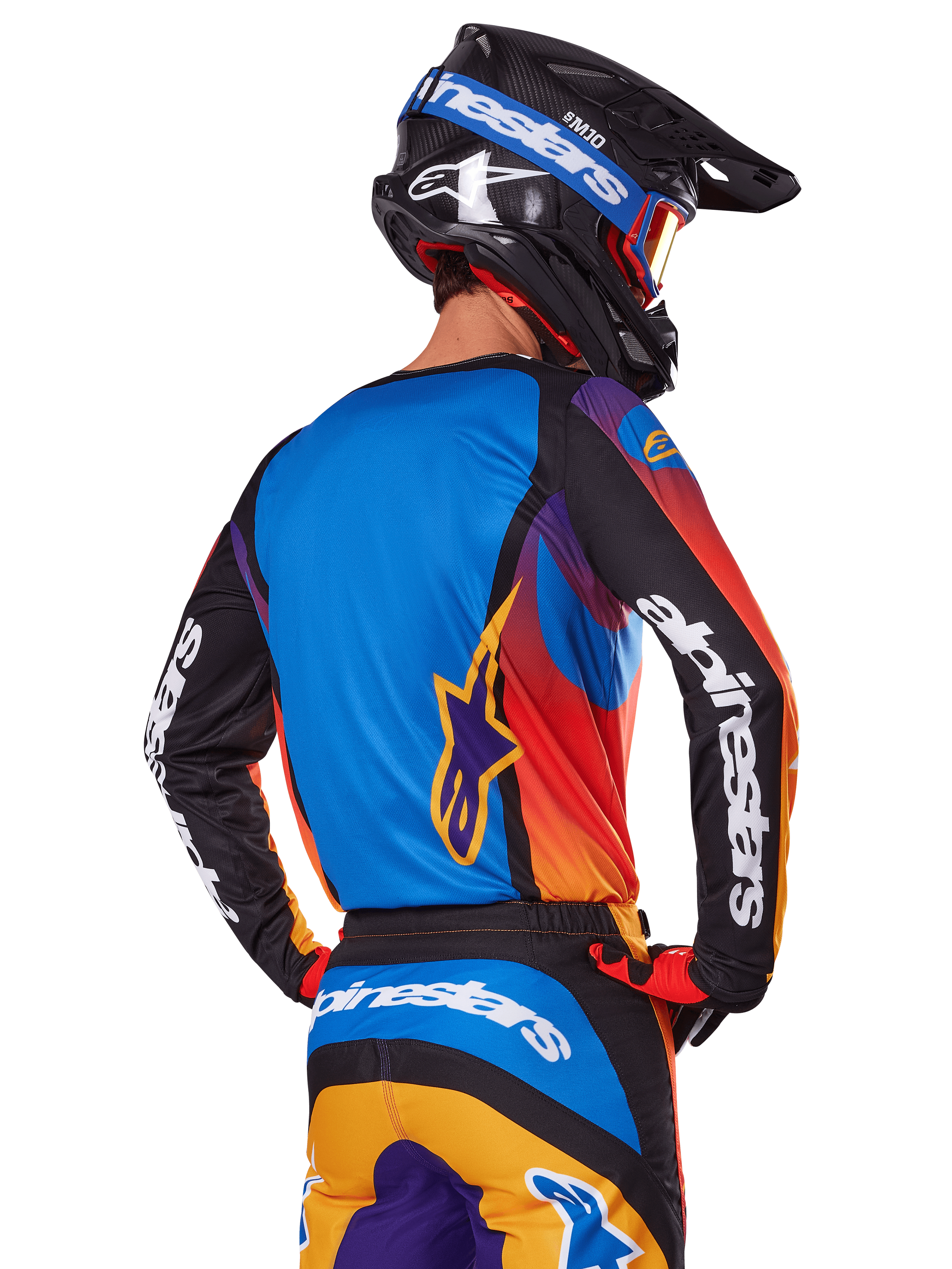 Motocrossförare sedd bakifrån som bär Alpinestars 2025 Fluid Wurx-utrustningsset, flerfärgad tröja med blå rygg och svarta ärmar med vita logotyper, matchande orangea och blå byxor, svart och blå hjälm med orangea goggles, koordinerad racingoutfit för terrängkörning.