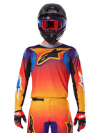 Alpinestars 2025 Fluid Wurx tröja, mångfärgad motocrosströja med orange och röd toning och lila accenter, bärs med matchande utrustning, svart hjälm med blå och röda goggles och röda handskar, med en stor svart stjärnlogotyp på bröstet.