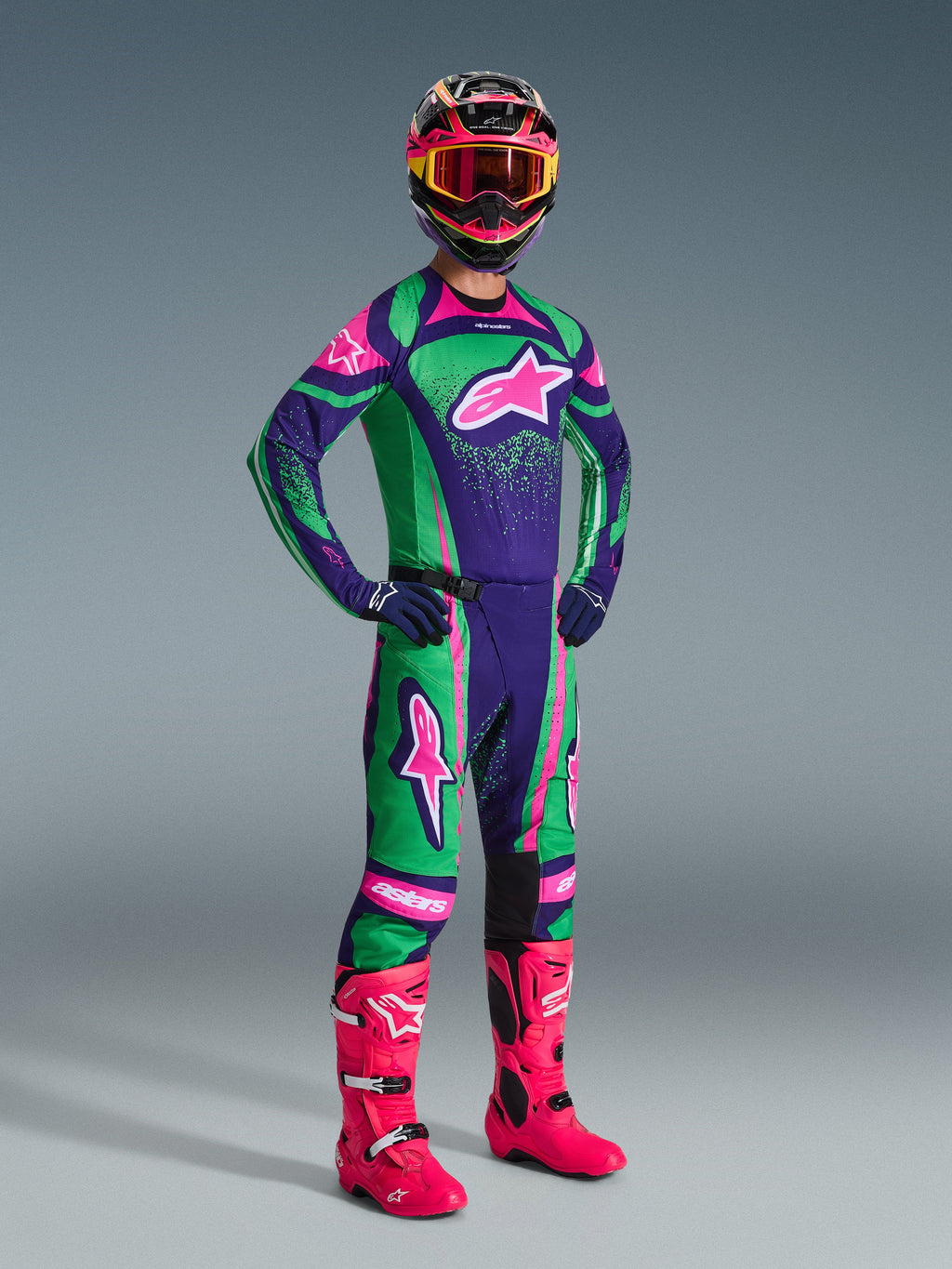 Motocross rider wearing the Alpinestars 2026 Techstar Nomur Gear Set in Deep Purple, Green, and Pink Fluo, featuring a matching jersey and pants, a colorful helmet with pink goggles, and vibrant pink boots, professional off-road racing equipment.