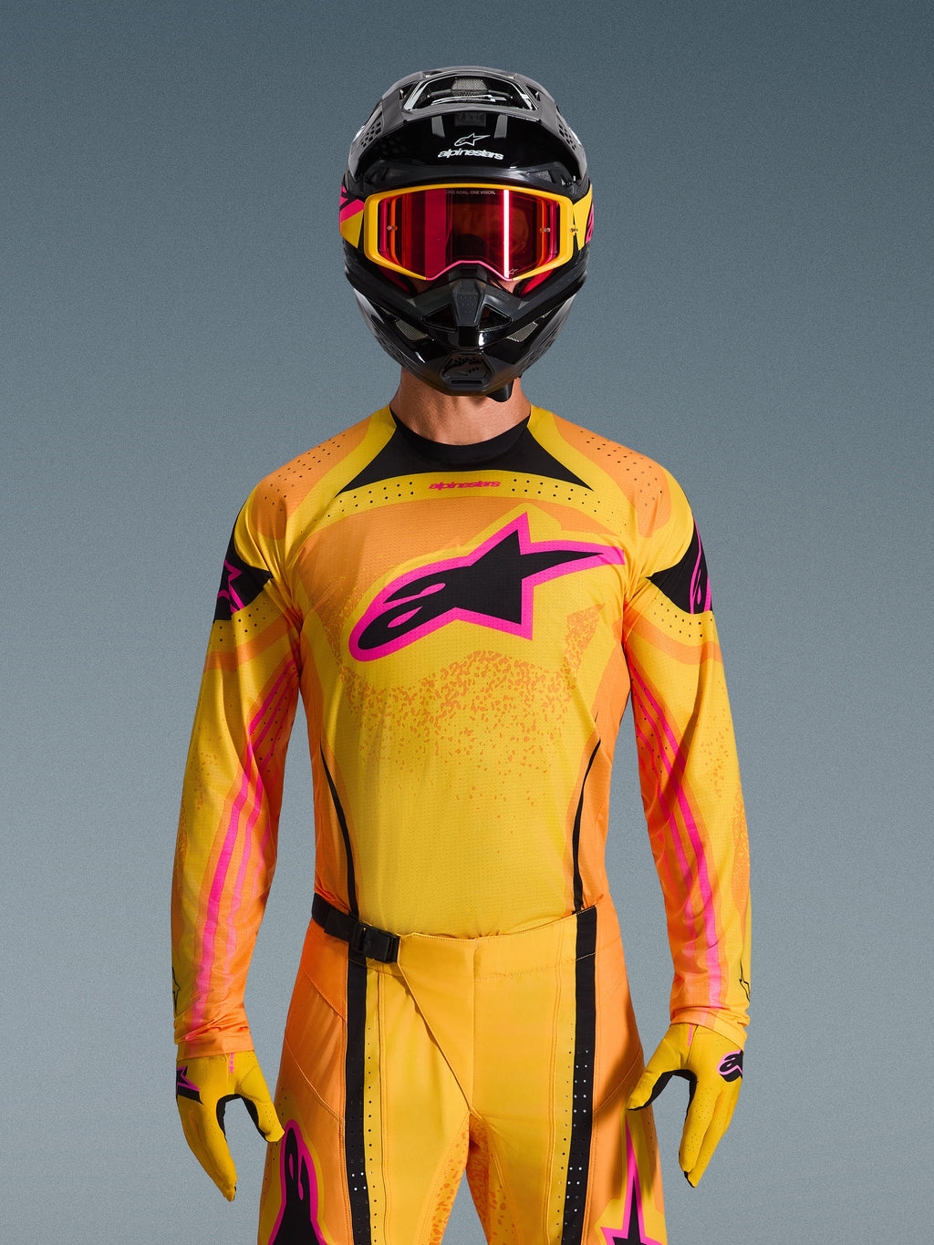 2026 Techstar Nomur-tröja i Orange Yellow Pink, buren av en motocrossförare med en matchande svart hjälm och rosa goggles, med en stor rosa Alpinestars-logotyp på bröstet, atletisk racingpassform för terrängprestanda.