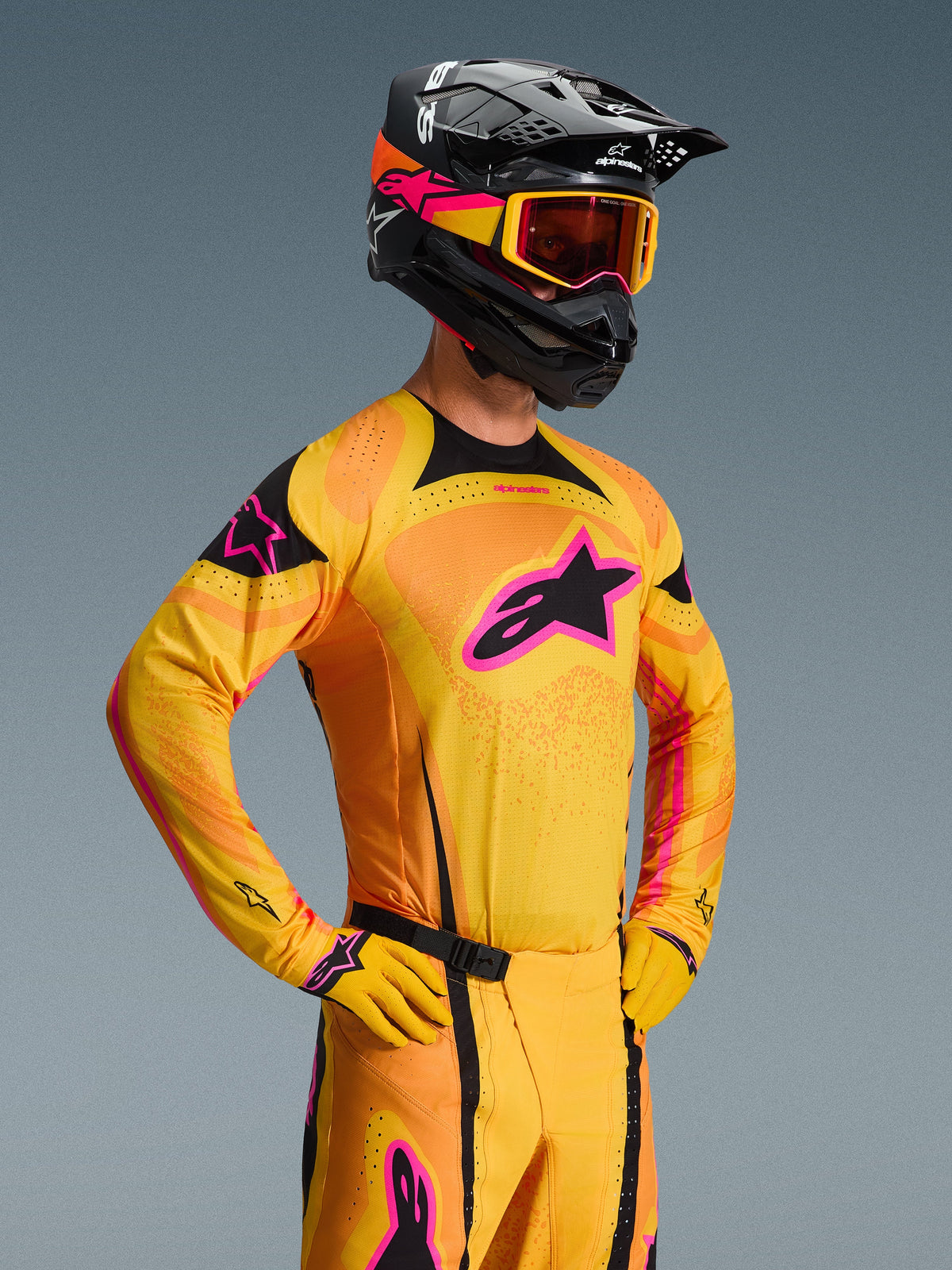Alpinestars 2026 Techstar Nomur Jersey, motocrosströja, orange, gul och rosa, bärs av en förare med matchande svart hjälm och goggles, med en stor rosa stjärnlogotyp på bröstet, svarta detaljer och laserskurna ventilationsdetaljer.