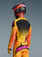 Alpinestars 2026 Techstar Nomur-tröja i orange, gult och rosa, vy bakifrån av en motocrossförare i komplett utrustning med matchande byxor och svart hjälm, livfull design med gula och rosa detaljer och svart ryggpanel, atletisk racingpassform för terrängkörning