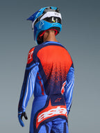 Motocrossförare sedd bakifrån iklädd 2026 Techstar Nomur-tröja, mörkblå med orangea och UCLA-blå detaljer, med en stor orange texturerad grafik på ryggen, matchad med koordinerande motocrossbyxor och en blå racinghjälm, professionell utrustning för terrängkörning.