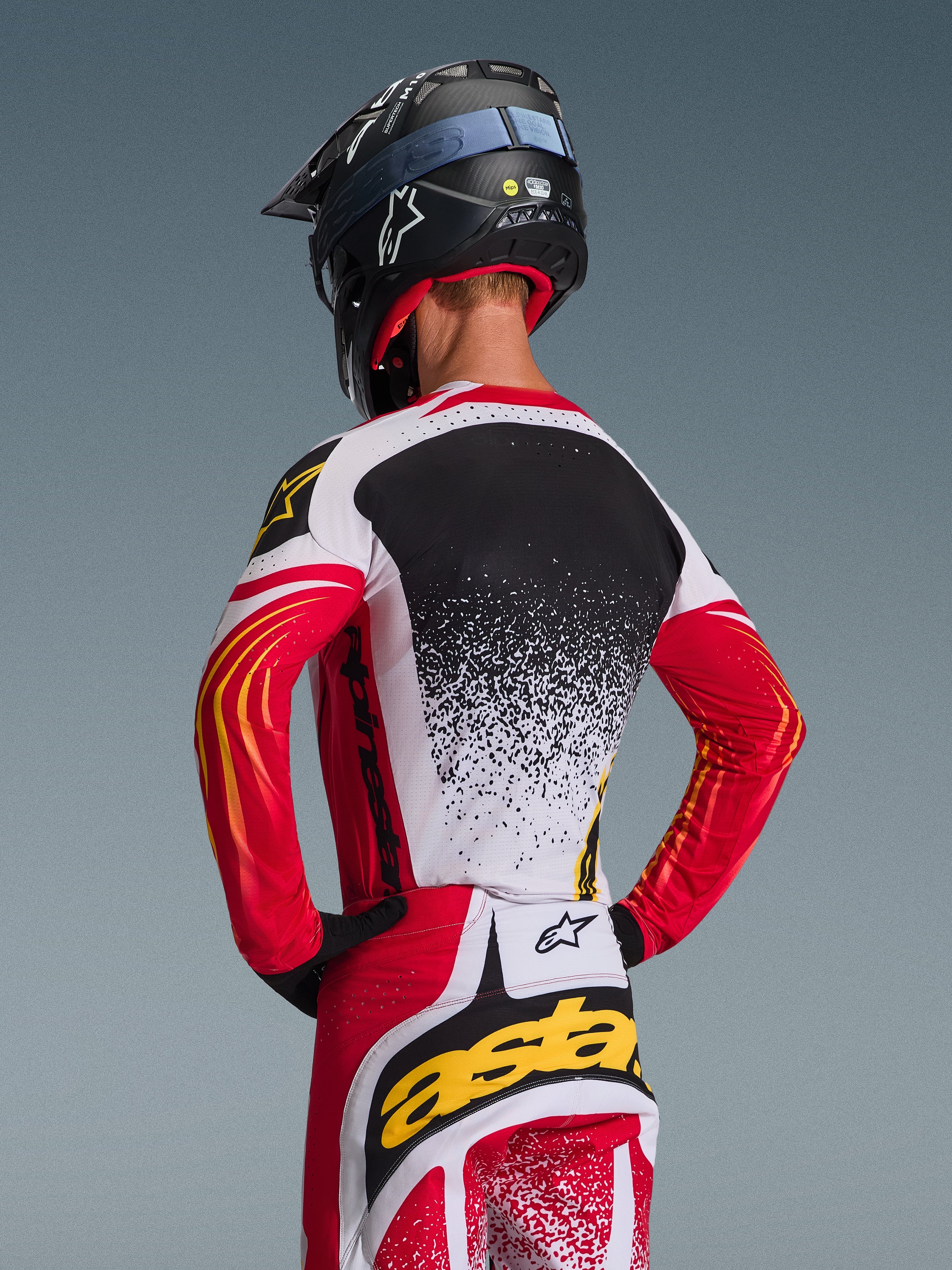 Alpinestars 2026 Techstar Nomur utrustningsset, motocrosströja och byxor, röd, vit och svart med gula detaljer, ryggvy av förare i svart hjälm, tekniska racingkläder med stänkgrafik och tydliga logotyper