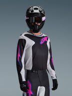 Motocross rider wearing the Alpinestars 2026 Fluid Grid Jersey, Black, Light Gray, and Purple, featuring large pink and purple star logos, paired with matching pants, black helmet, and goggles, complete off-road racing gear set