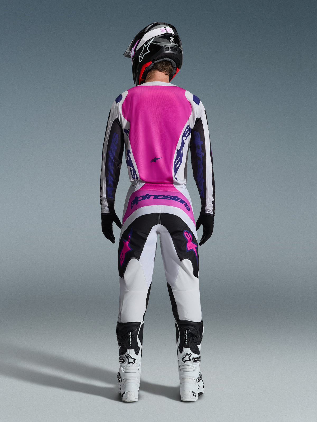 Motocrossförare sedd bakifrån i Alpinestars 2026 Fluid Grid-utrustningsset, med en klarrosa och vit tröja med svarta ärmar, matchande vita byxor med svarta och rosa paneler, lila Alpinestars-branding, vita och svarta racingstövlar och en koordinerad hjälm, designad för off-road-prestanda.