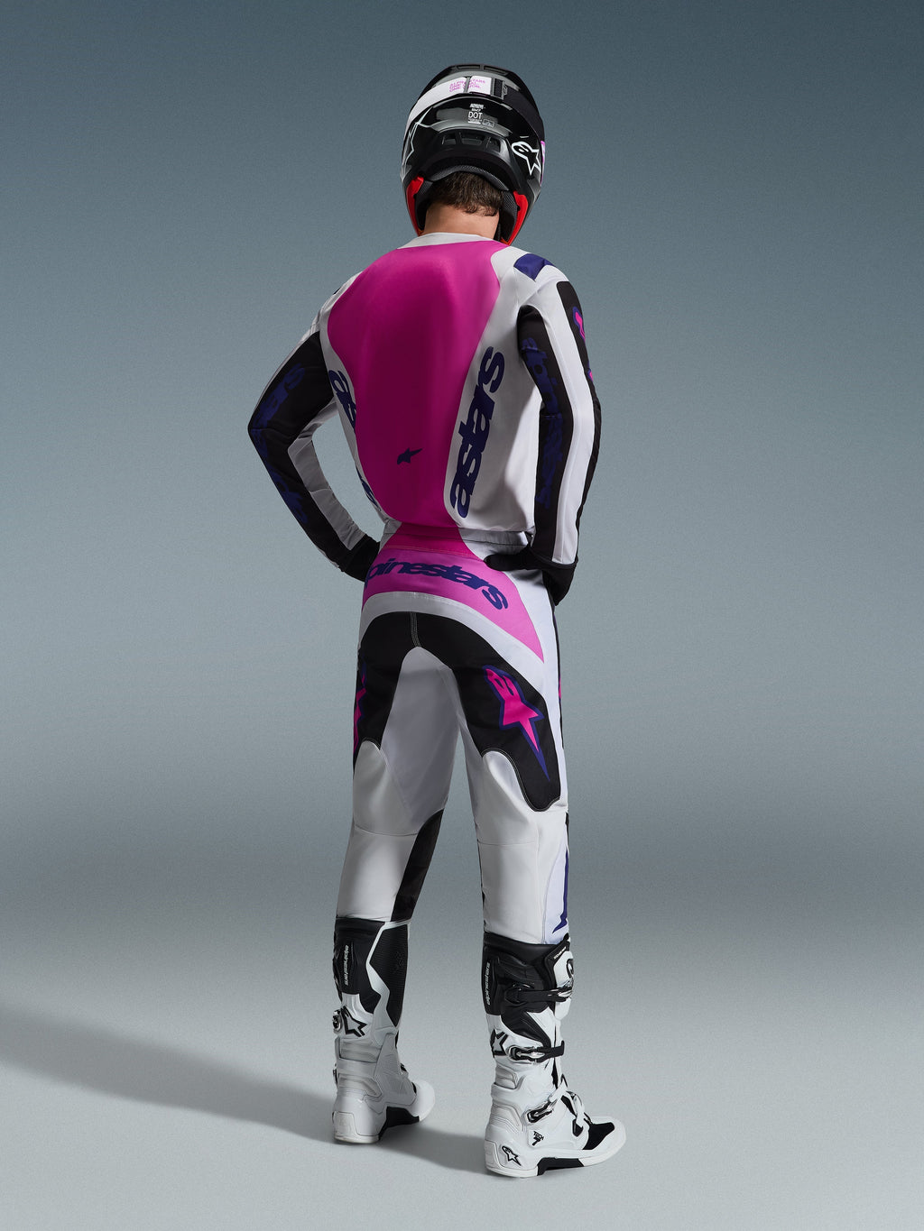 Alpinestars 2026 Fluid Grid Gear Set, Motocross rider seen from the back, white jersey with a pink back panel and purple branding, matching white and black pants with pink accents, white and black boots, and a black helmet, coordinated off-road racing outfit.