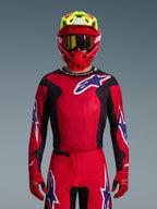 Motocrossförare i Alpinestars 2026 Fluid Grid-utrustningsset, röd tröja med svarta sidopaneler och vit stjärnlogotyp, matchande röda byxor och handskar, röd och gul hjälm med guldtonade goggles, högpresterande racingutrustning för terrängkörning