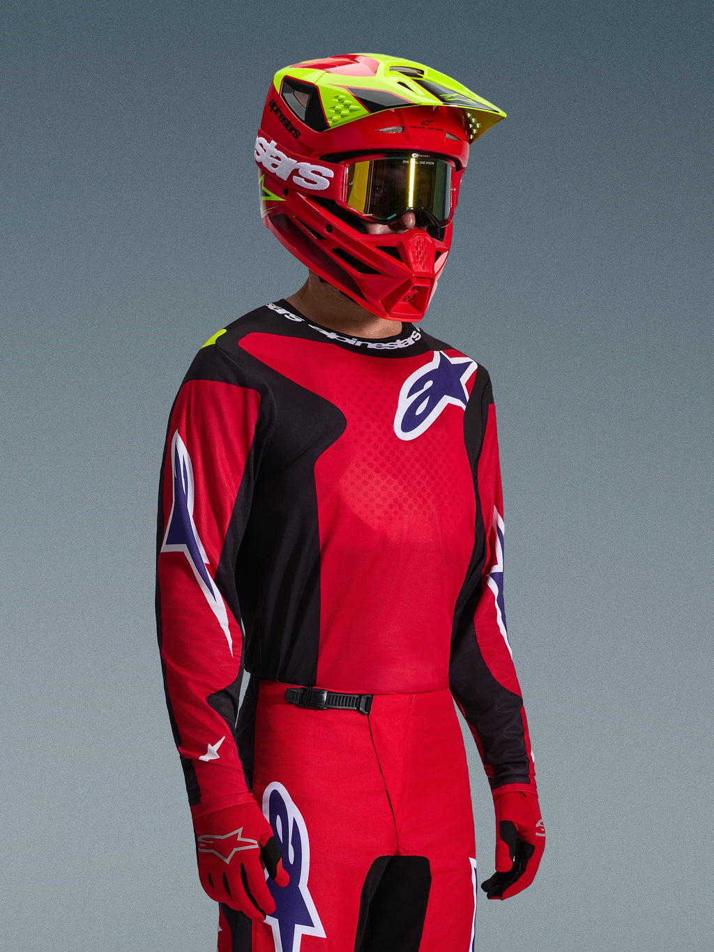 Motocrossförare iklädd Alpinestars 2026 Fluid Grid-utrustningsset, röd och svart tröja med vita och blå logotypdetaljer, matchande röda och svarta byxor, röd hjälm med gula detaljer och guldiga goggles, koordinerad offroad-racingutrustning