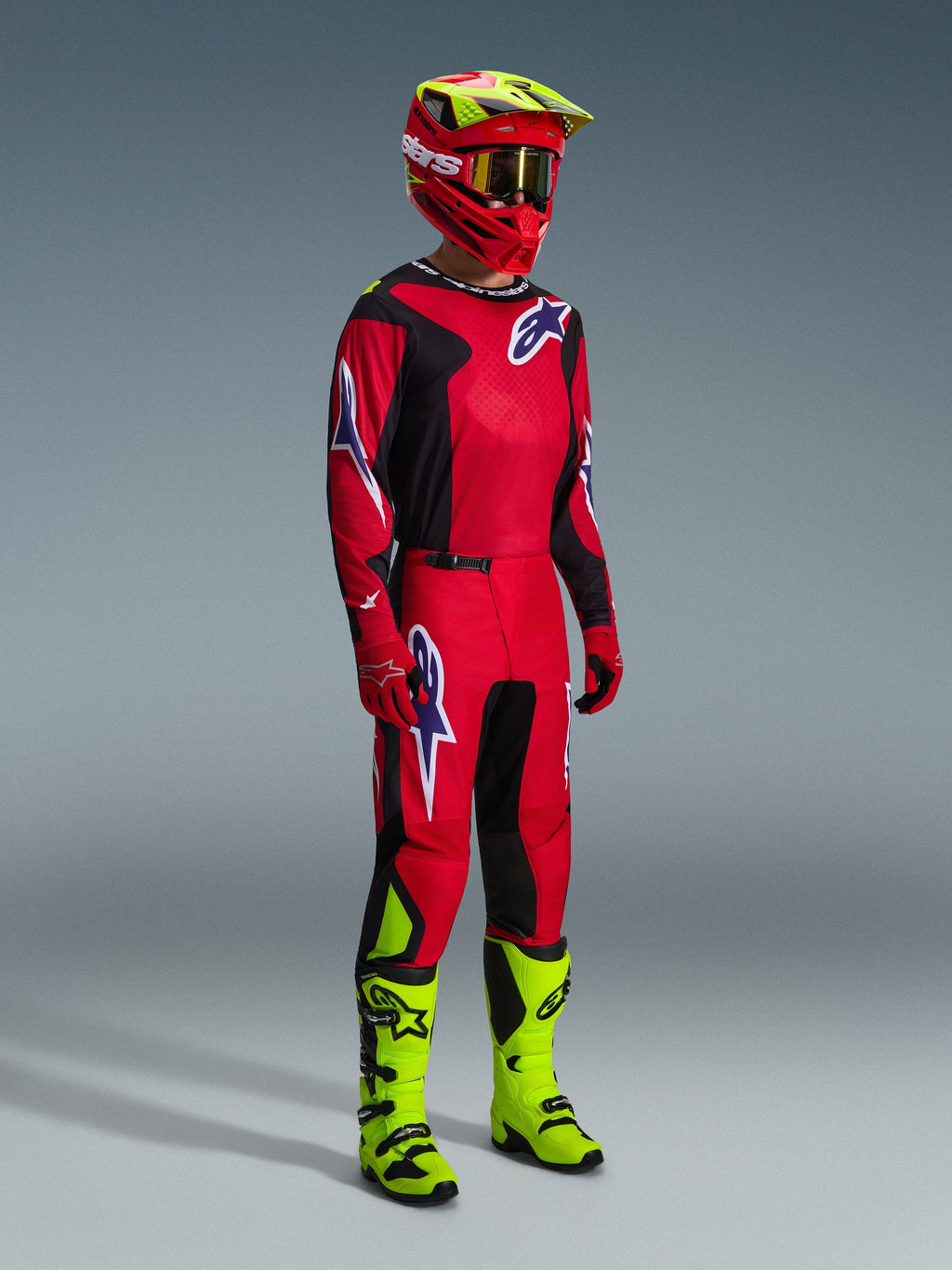 Motocrossförare iklädd Alpinestars 2026 Fluid Grid-utrustningsset i rött, svart och lila, med matchande tröja och byxor med lila logotyper, en röd och neongul hjälm och neongula stövlar för en komplett outfit för terrängkörning.