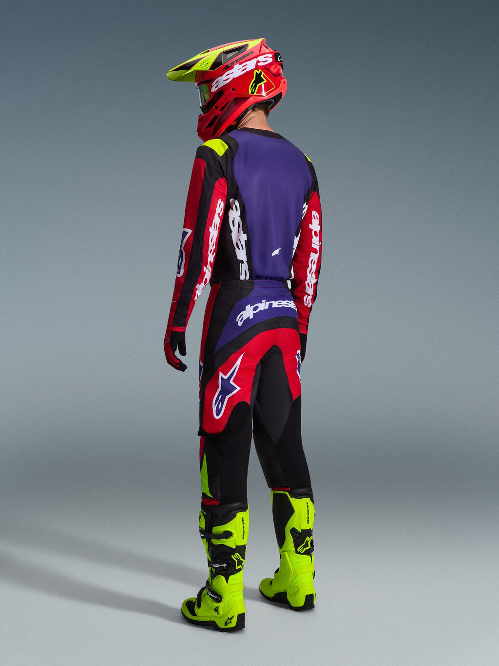 Motocrossförare sedd bakifrån i Alpinestars 2026 Fluid Grid-utrustningsset, röd, svart och lila tröja med vit logotyp, röda och svarta byxor med lila detaljer, röd och neongul hjälm, starkt neongula stövlar, komplett offroad-racingutrustning