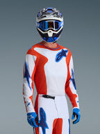 Motocross rider wearing the Alpinestars 2026 Fluid Grid Gear Set, orange and white jersey with blue star logos, white and orange pants with blue accents, blue gloves, blue and white helmet with blue goggles, coordinated athletic racing gear for off-road performance