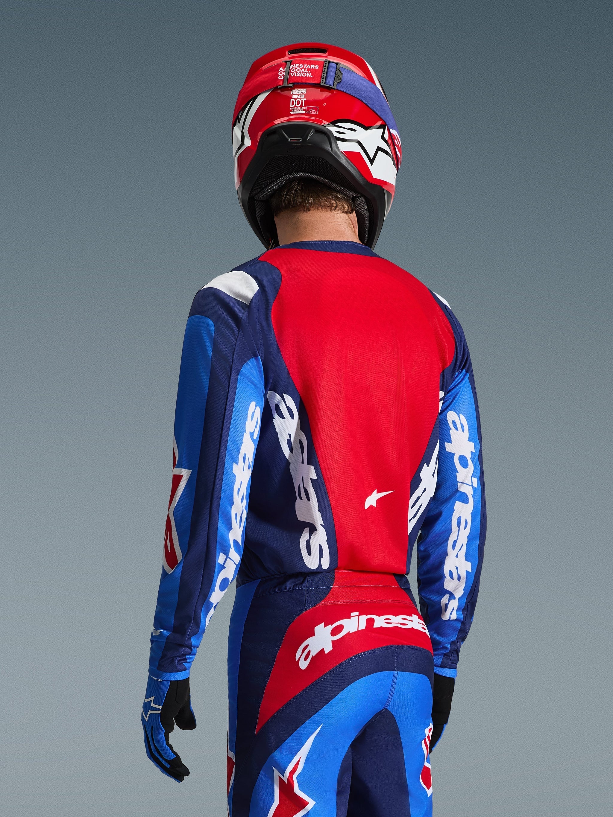 Motocrossförare sedd bakifrån i Alpinestars 2026 Fluid Grid Jersey i blått och rött, med tydliga vita logotyper på ärmarna och ryggen, kombinerat med matchande motocrossbyxor och en röd hjälm för en komplett racinglook för terrängkörning