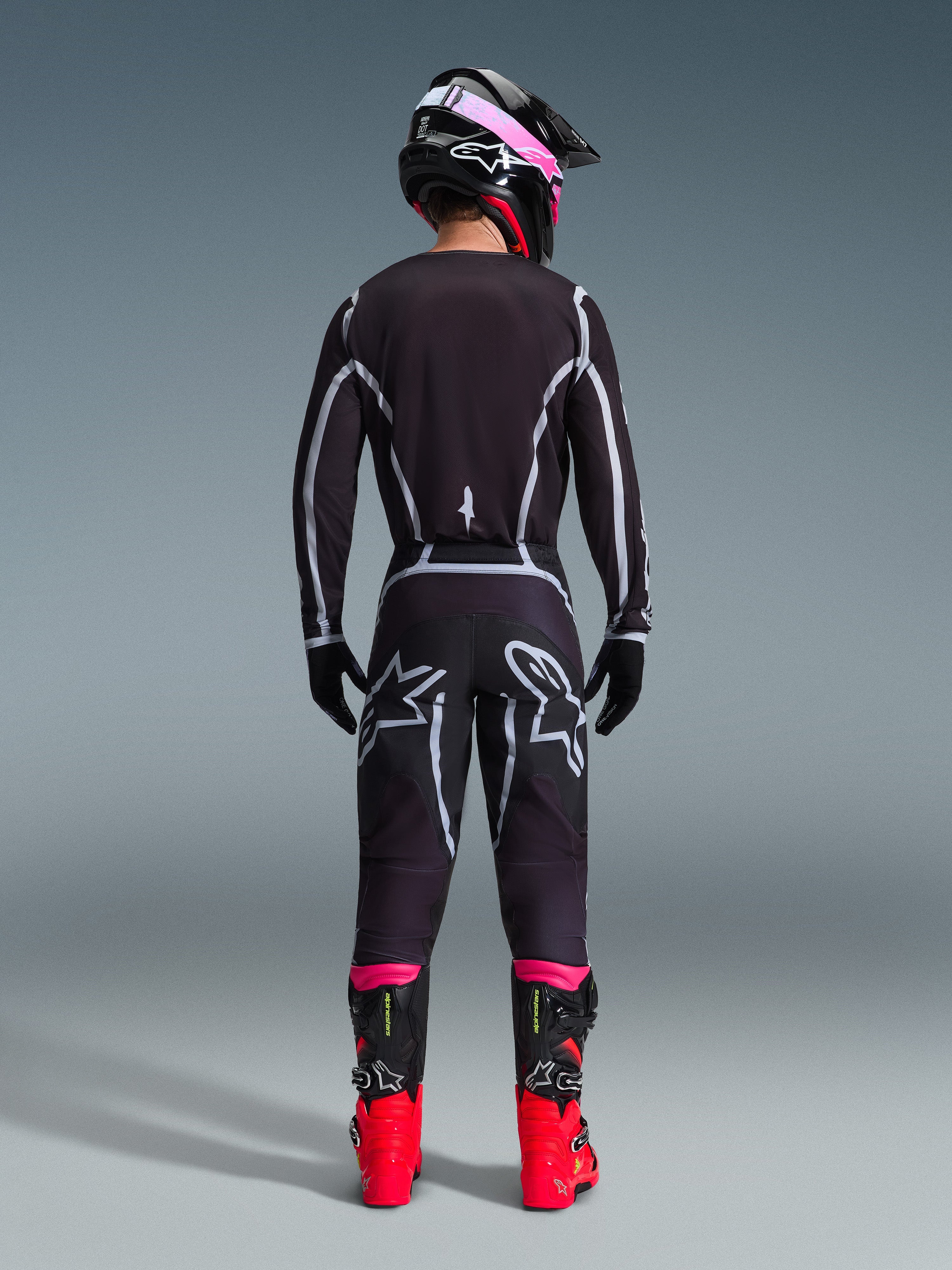 Motocross rider seen from behind wearing the Alpinestars 2026 Fluid Apex Gear Set, black jersey and pants with grey geometric lines and star logos, black helmet with pink accents, bright red and black boots, coordinated motocross racing gear