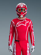 Motocrossförare i Alpinestars 2026 Fluid Apex utrustningsset, röd tröja med stor vit stjärnlogotyp och vita linjer, matchande röda byxor med vita detaljer, röd och vit hjälm med lila inslag och orangefärgade goggles, vita och svarta handskar, koordinerad outfit för terrängkörning