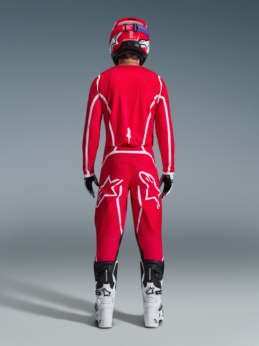 Vy bakifrån av en motocrossförare iklädd Alpinestars 2026 Fluid Apex-utrustningsset, klarröd tröja och byxor med vita ränder och logotyper, vita och svarta stövlar och en röd hjälm, professionell racingutrustning för terrängkörning