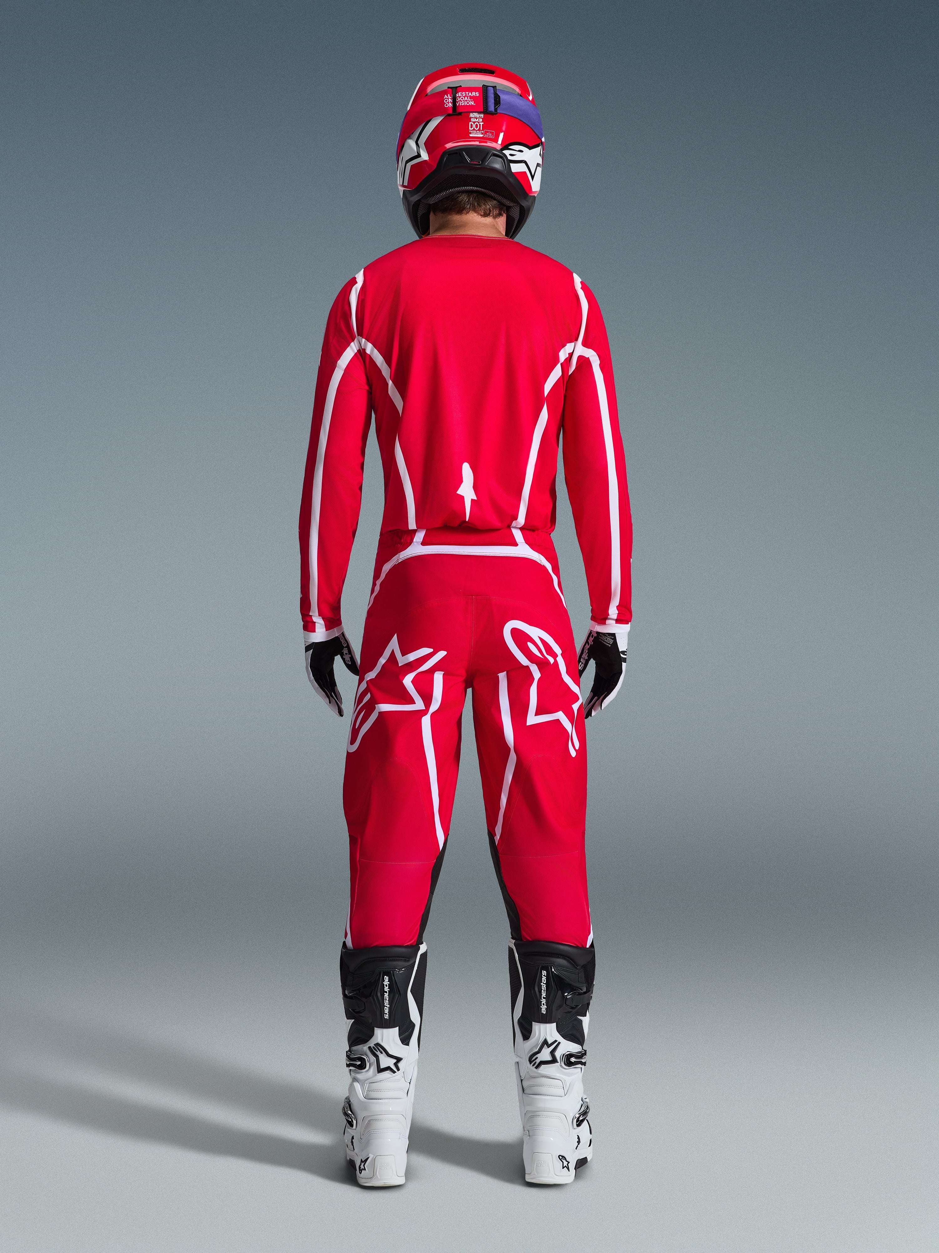 Vy bakifrån av en motocrossförare iklädd Alpinestars 2026 Fluid Apex-utrustningsset, klarröd tröja och byxor med vita ränder och logotyper, vita och svarta stövlar och en röd hjälm, professionell racingutrustning för terrängkörning