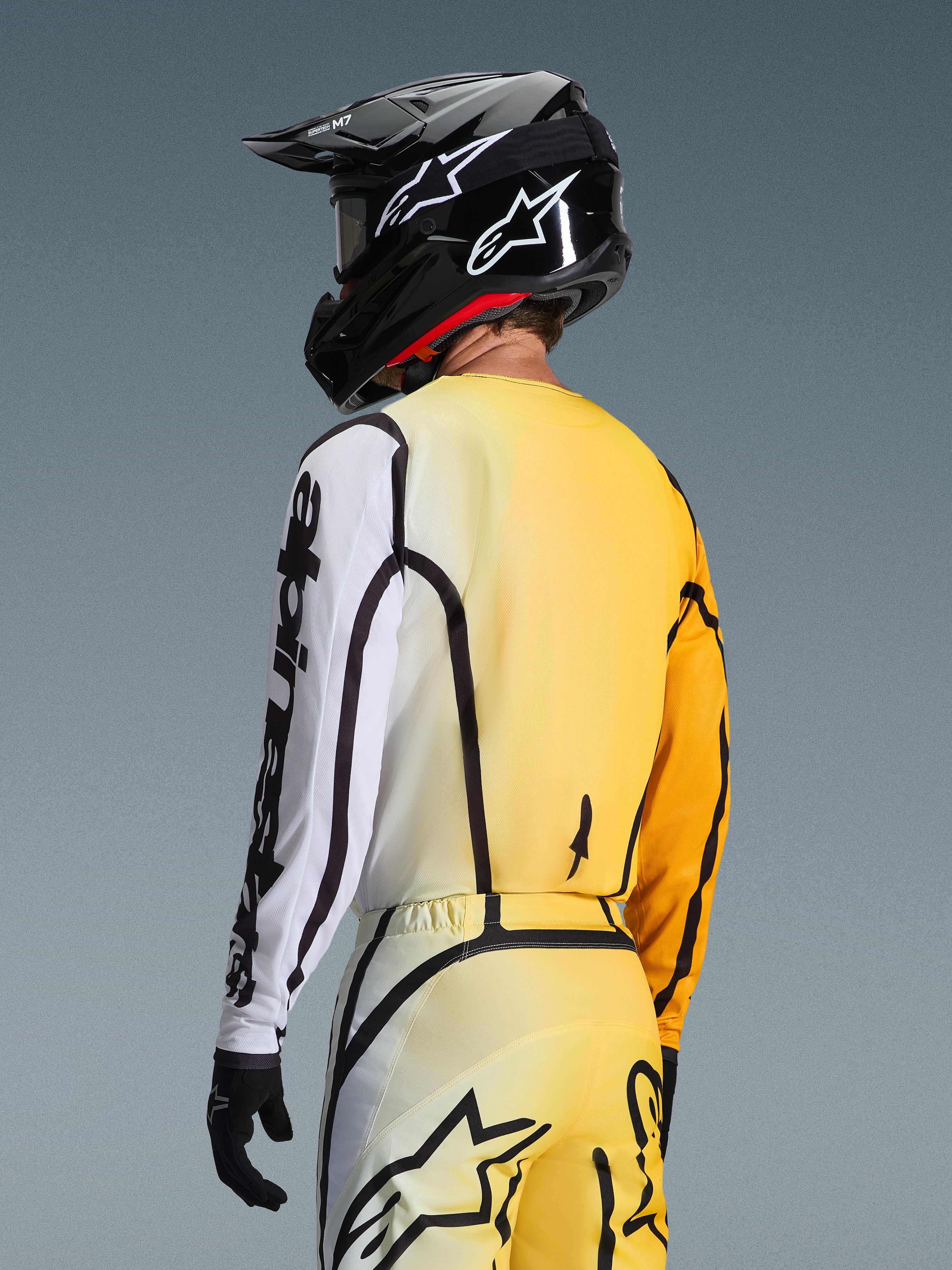 Bakre trekvartsvy av en motocrossförare som bär 2026 Fluid Apex-tröjan i färgen Yellow Black, med en tonad design från gult till vitt, tydlig svart Alpinestars-logotyp på ärmen, matchande byxor och en svart motocrosshjälm.