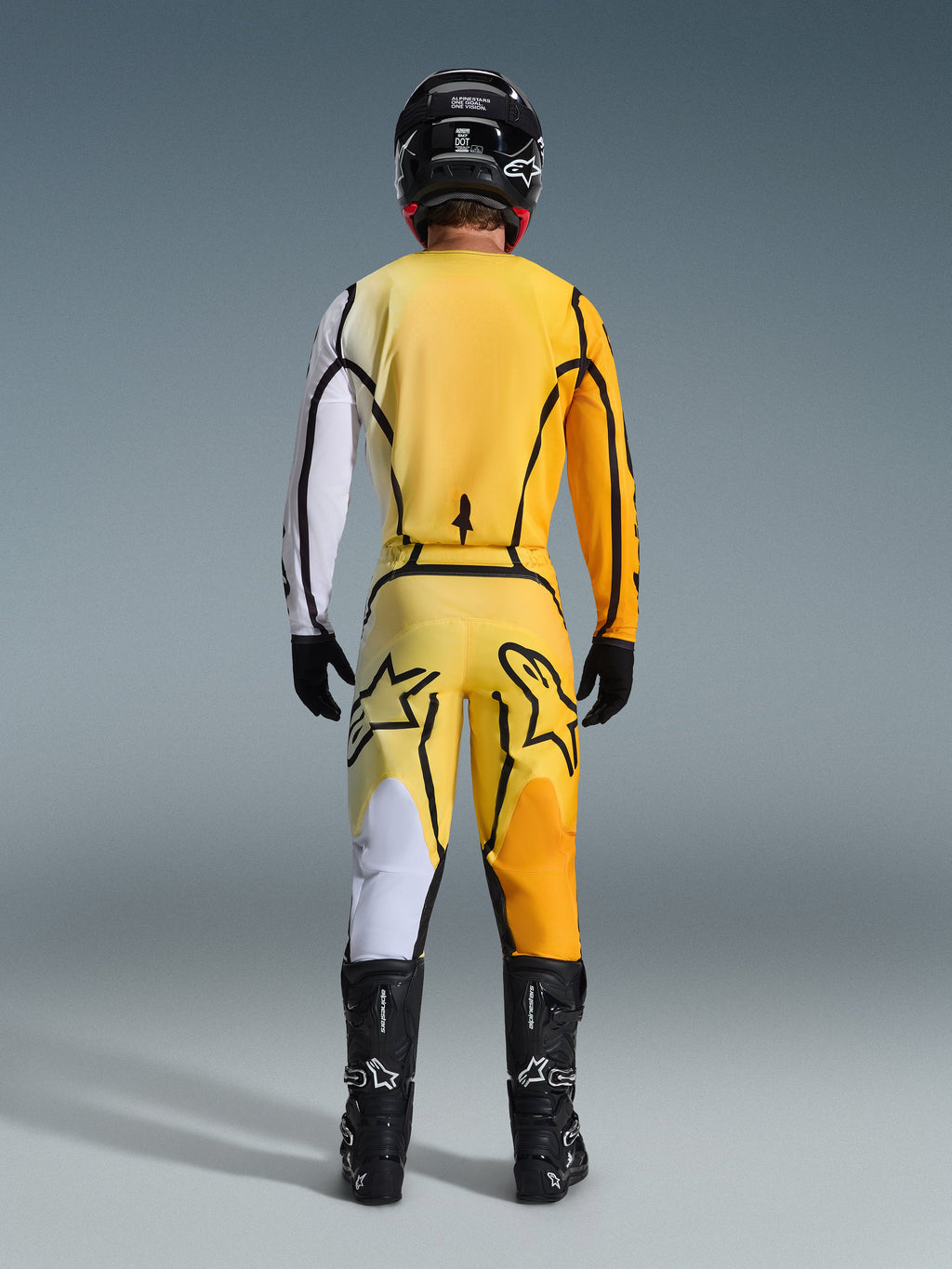 Motocrossförare sedd bakifrån bär Alpinestars 2026 Fluid Apex-tröja och matchande byxor i Yellow Black, med framträdande färgblockspaneler i vitt, gult och orange, kombinerat med en svart hjälm och motocross-stövlar.