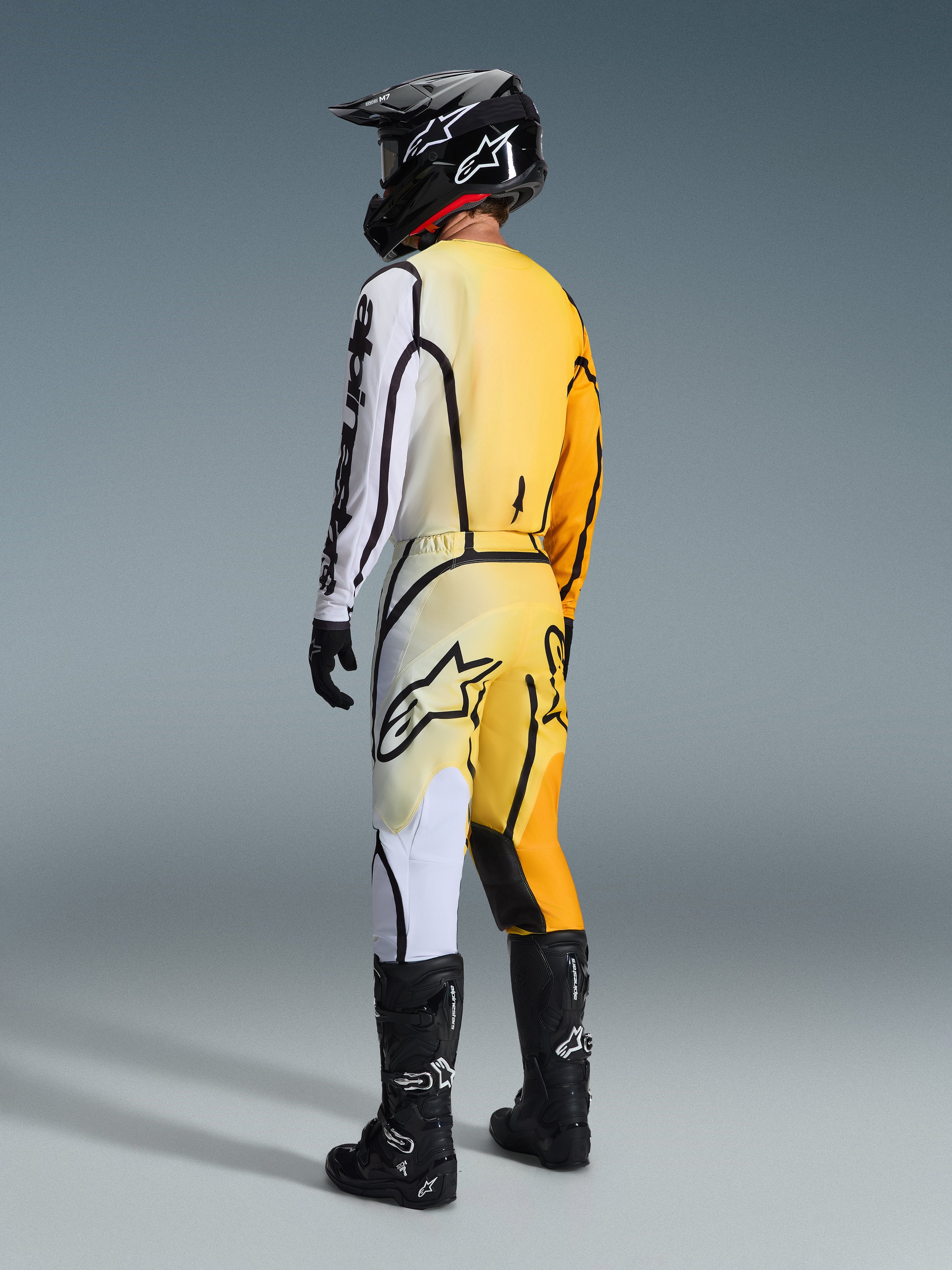 Motocrossförare sedd bakifrån iförd Alpinestars 2026 Fluid Apex-tröja och matchande byxor, gult och svart utrustningsset med toningar från gult till vitt och kraftfulla svarta grafiska linjer, kombinerat med svart hjälm, svarta och vita handskar och svarta motocross-stövlar.