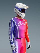 Alpinestars 2026 Fluid Apex tröja, motocrosströja, livfull gradient i lila, rosa och orange, bärs med matchande byxor, vit hjälm med blå goggles och vita handskar, atletisk design för offroad-prestanda och rörlighet