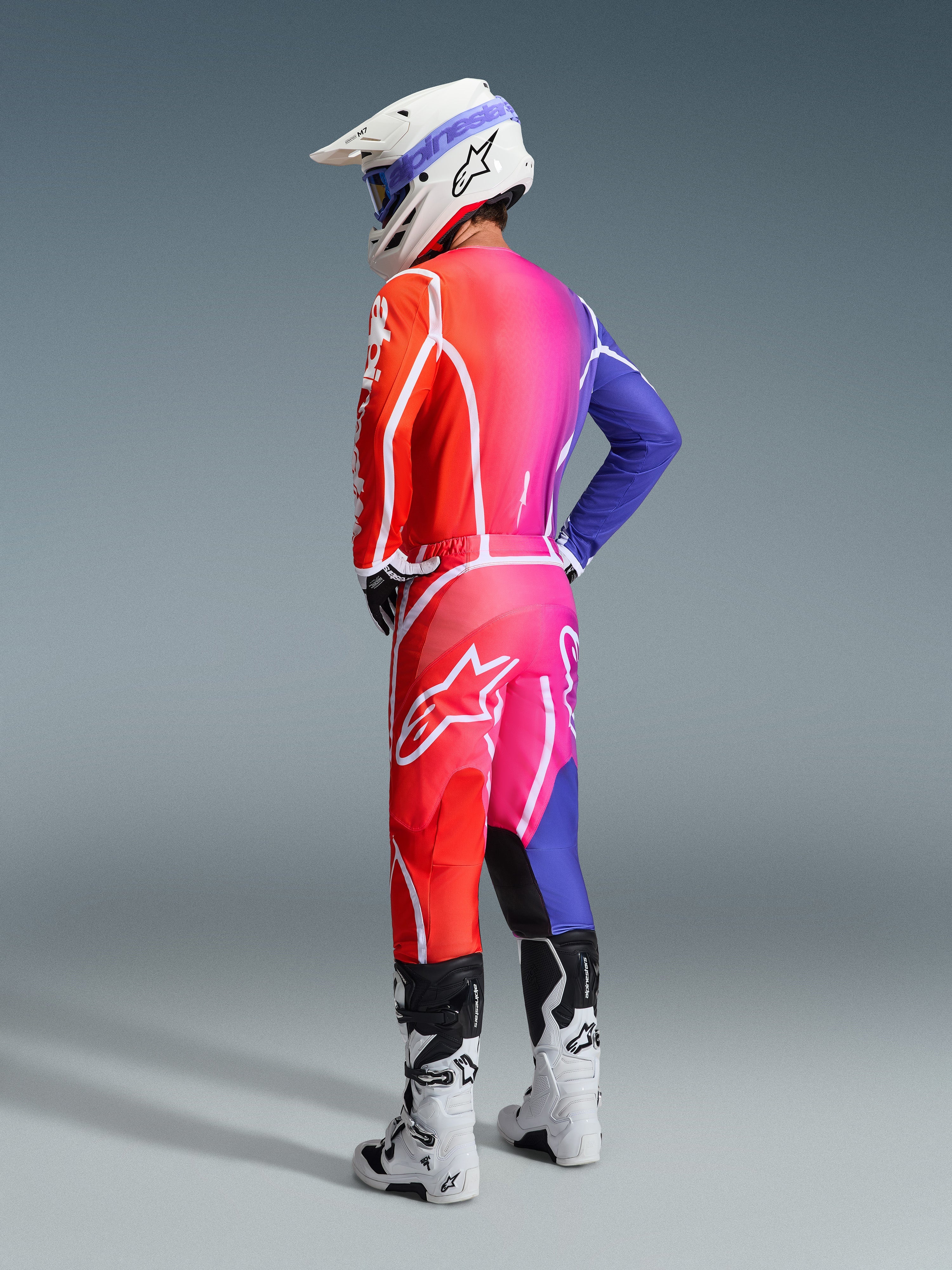 Motocrossförare som bär Alpinestars 2026 Fluid Apex-utrustningsset, livfull färgtoning från orange till rosa och lila med framträdande vit logotyp och stjärnsymboler, kombinerat med en vit hjälm, blå goggles och vita stövlar, sedd ur en trekvartsvinkel bakifrån i studiomiljö.