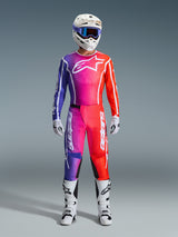 Motocrossförare i komplett Alpinestars 2026 Fluid Apex racingutrustning, med tröja och byxor i lila, rosa och orange gradient, vit Alpinestars-logotyp på bröstet, vit hjälm med blå goggles och vita stövlar, designad för offroad-prestanda.